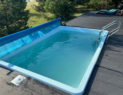 Композитный бассейн с металлической лестницей и деревянной террасой на фоне хвойных деревьев